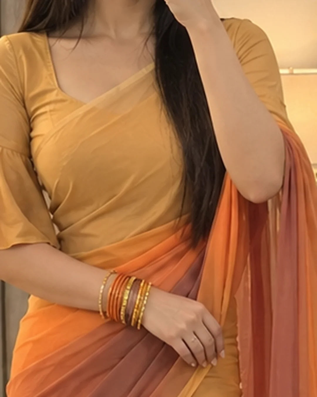 A woman wearing a mustard yellow and orange gradient saree with a matching short-sleeve blouse, accessorized with gold and red bangles.