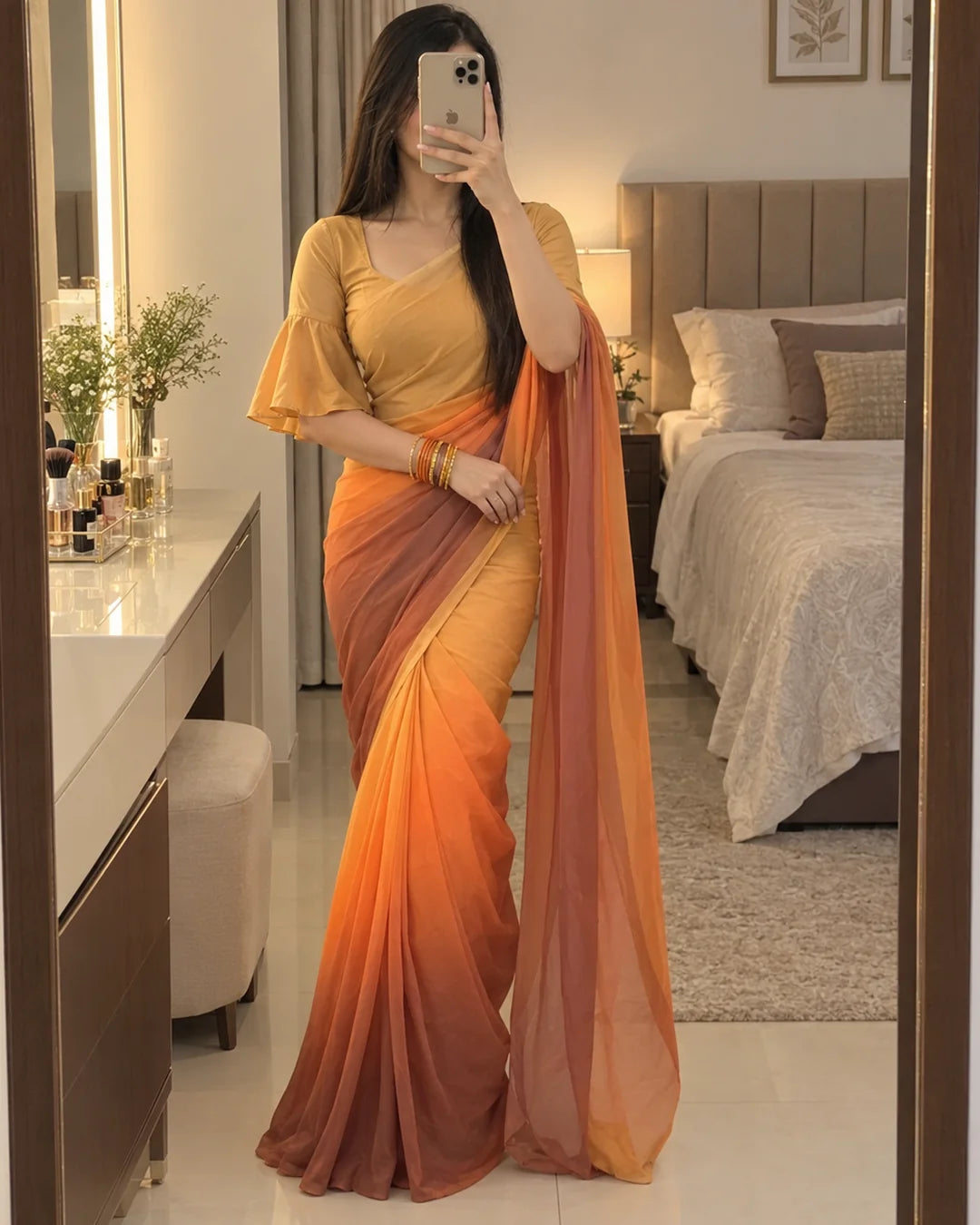 A woman takes a mirror selfie in a bedroom wearing a yellow and orange ombre gradient saree with a matching yellow blouse.